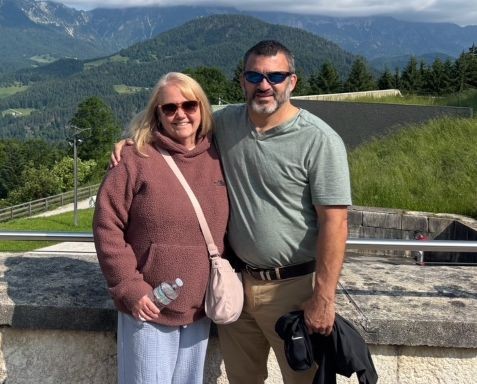Smiling couple posing outdoors on a stone railing with green hills and mountains in the background; woman holds a water bottle and wears a pink hoodie.