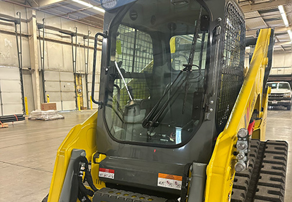 Yellow compact track loader with enclosed cab inside a warehouse or service bay, front view showing crawler tracks and protective cages.